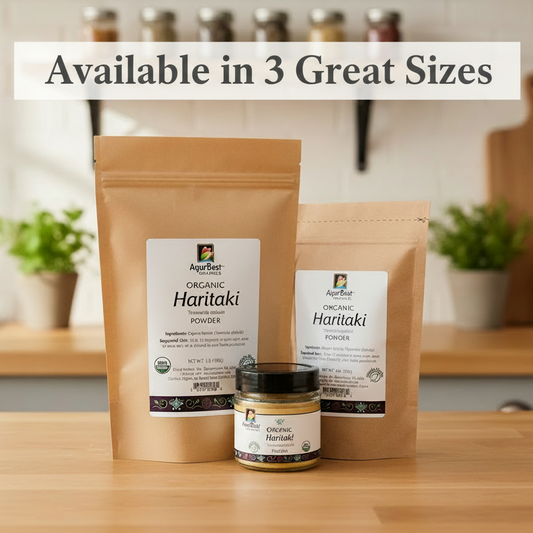 Three sizes of AyurBest Organic Haritaki Powder packaging on a wooden surface with plants in the background.