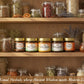 Six jars of Nityam "Daily" Superfood spreads on a shelf with herbs and foods in pantry.